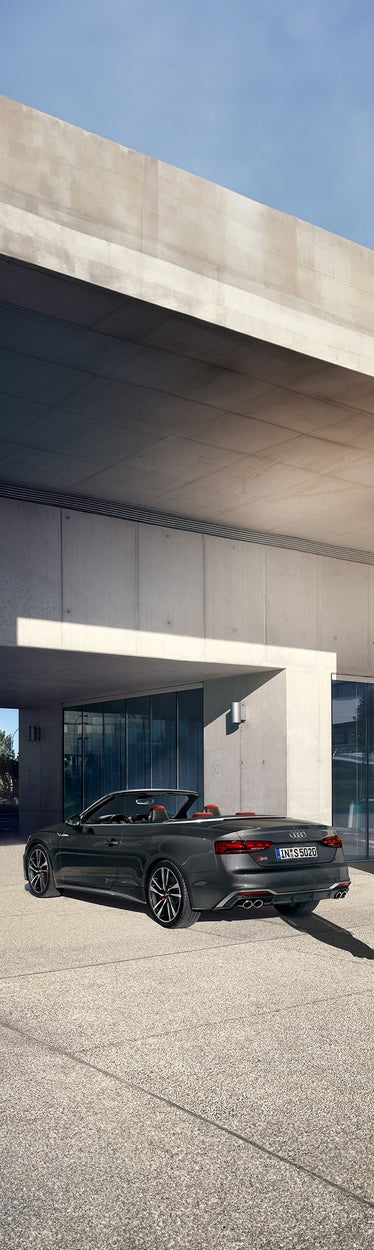 Dunkelgraues Audi S5 Cabriolet mit offenem Verdeck steht vor moderner Betonarchitektur, Frau in heller Kleidung geht vorbei.