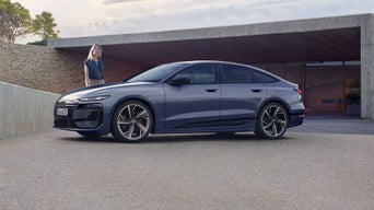 Der A6 Sportback e-tron steht im Seitenprofil vor dem Carport eines Hauses. Daneben steht eine junge Frau.