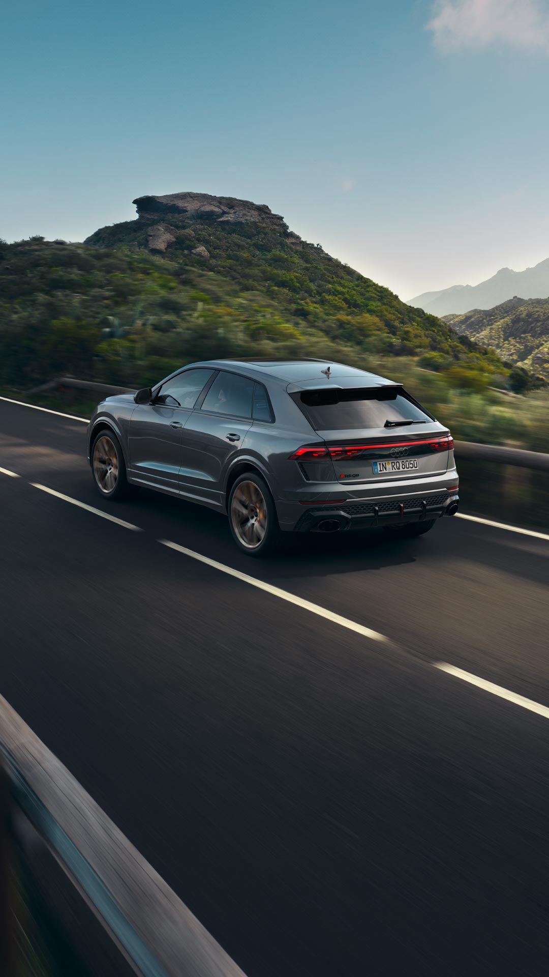 Ein grauer Audi RS Q8 SUV fährt auf einer kurvigen Straße durch grüne Berglandschaft.