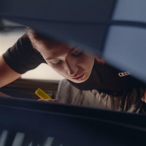 Woman in a workshop, intently focused on a task, holding a yellow tool. Dim lighting and serious expression convey concentration and precision.
