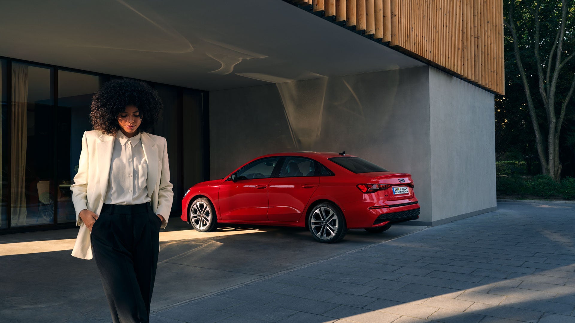 Eine Frau im weißen Blazer schaut nach unten, im Hintergrund steht ein roter Audi vor einem modernen Gebäude.