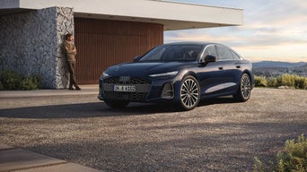 Dunkelblaue Audi A6 Limousine steht vor einem modernen Haus, ein Mann lehnt entspannt an einer Steinwand im Hintergrund.