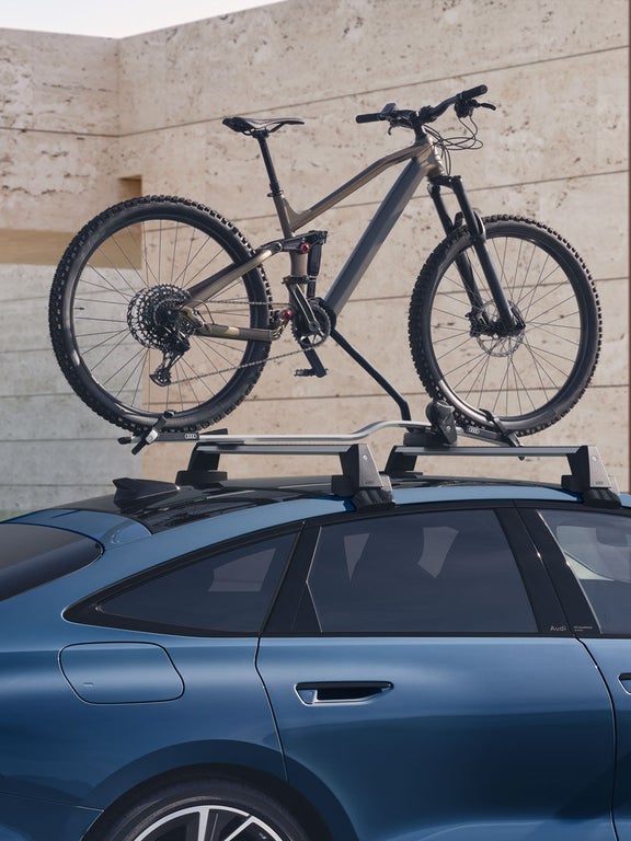 Blauer Audi A6 Sportback e-tron mit montiertem Mountainbike auf dem Dachträger vor moderner Hausfassade.