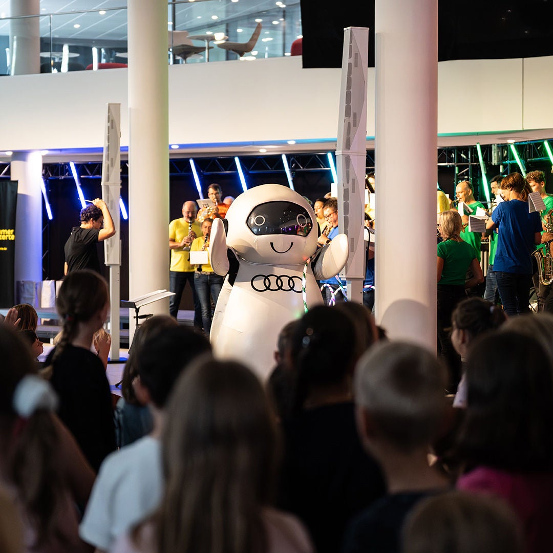 Weißes Audi Robotermaskottchen ADUI zwischen Publikum und Blasorchester.