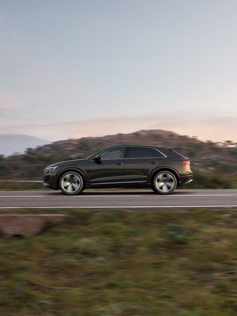 Ein schwarzer Audi Q8 SUV TFSI e fährt auf einer Landstraße vor hügeliger Landschaft bei Tageslicht.
