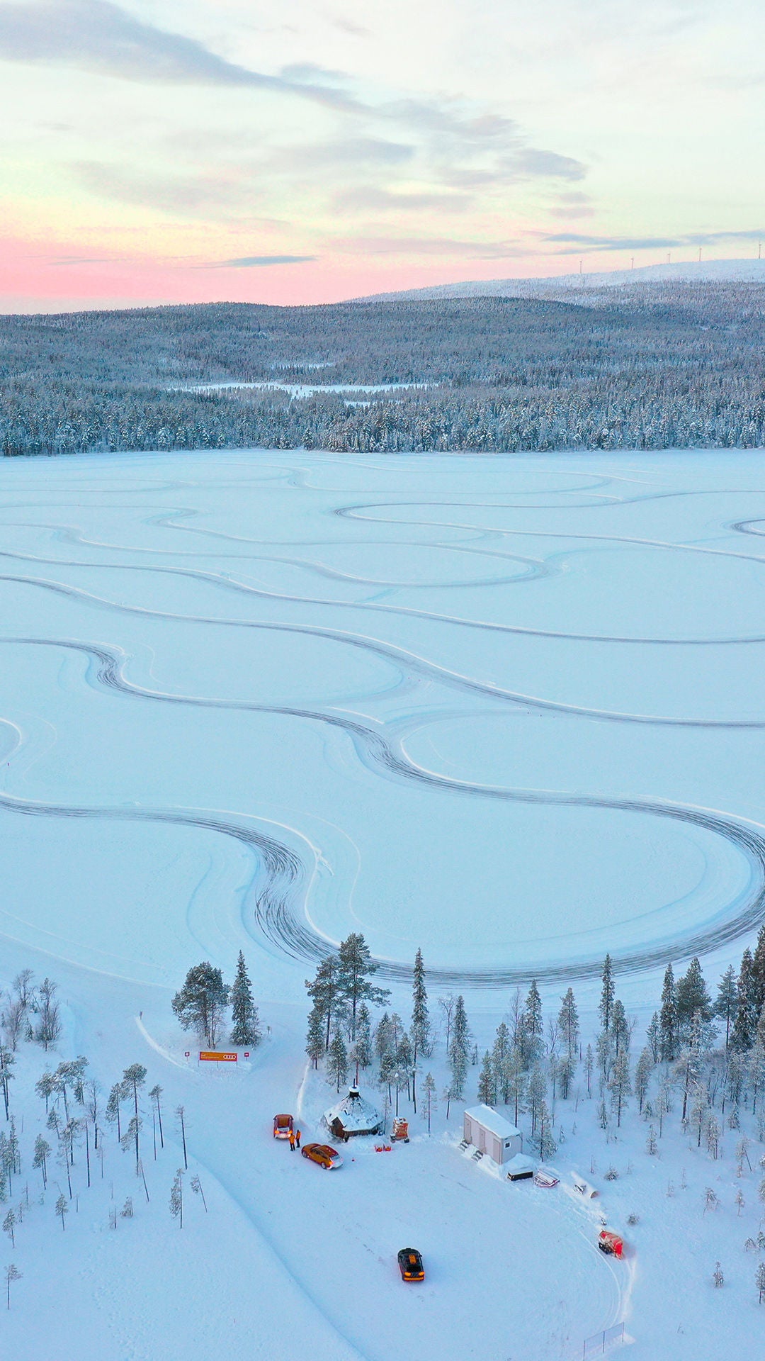 Verschneite Landschaft mit geschwungenen Fahrspuren auf einem zugefrorenen See, umgeben von Wald und winterlichem Himmel.