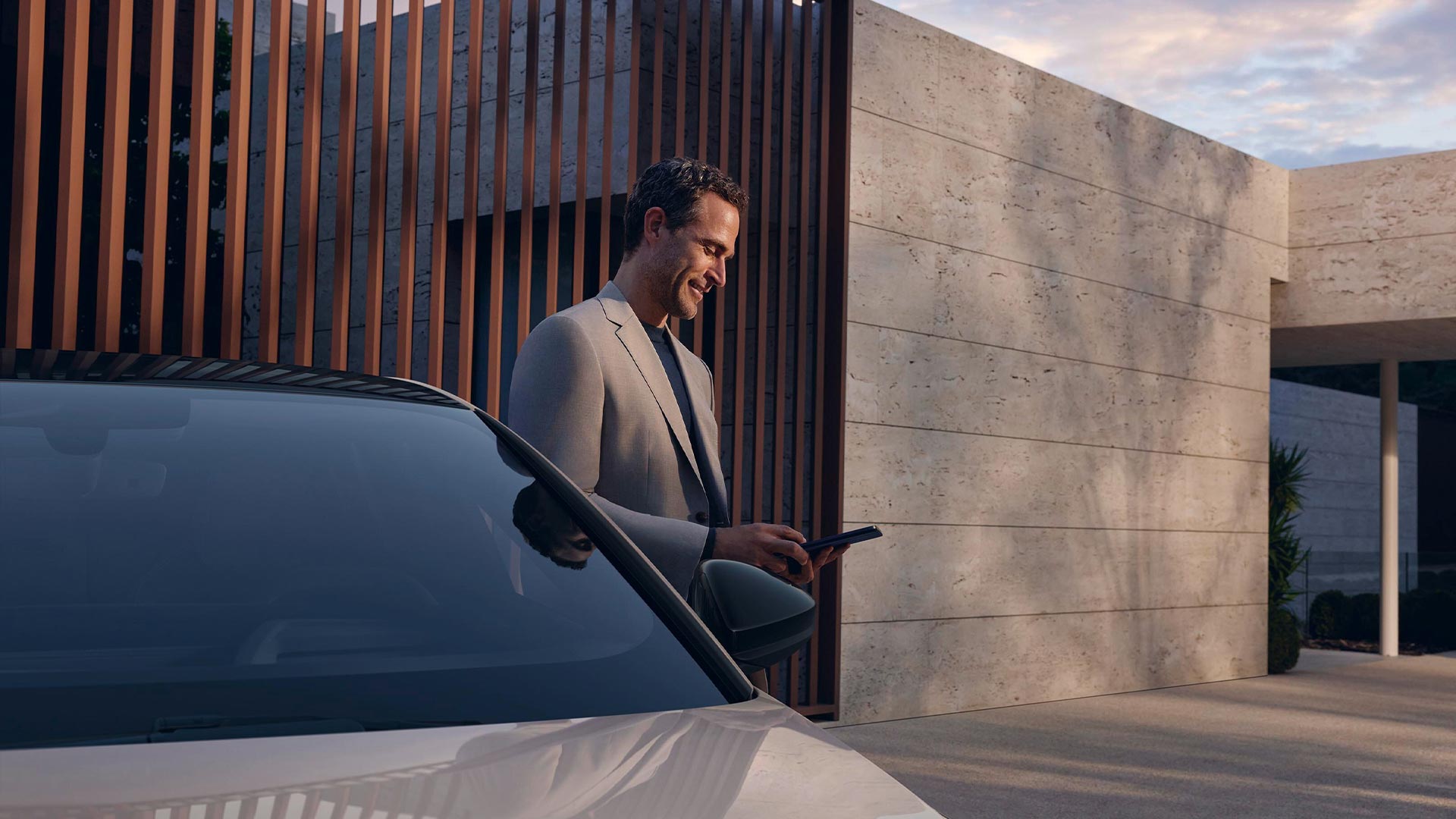 Ein Mann in grauem Anzug steht vor einem Haus und schaut lächelnd auf sein Smartphone neben einem geparkten Audi.