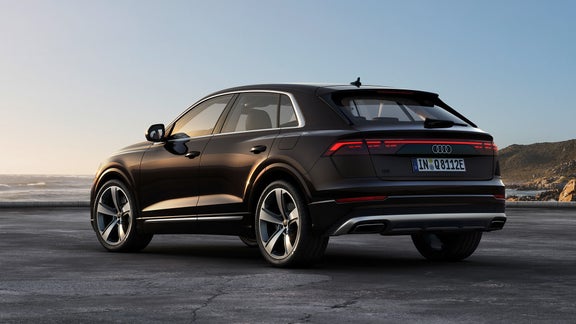 Ein schwarzer Audi Q8 TFSI e steht mit seiner eleganten Silhouette vor einer malerischen Küstenlandschaft. Die untergehende Sonne spiegelt sich auf der Karosserie, während das rote LED-Leuchtenband am Heck markant hervorsticht.