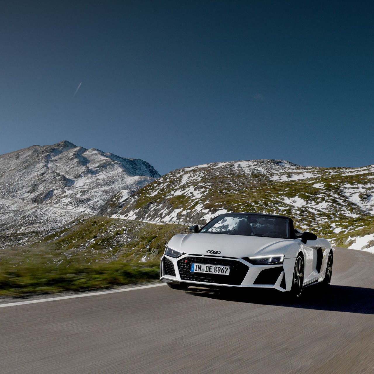 Weißer Audi R8 Spyder fährt auf einer Bergstraße mit schneebedeckter Landschaft im Hintergrund.