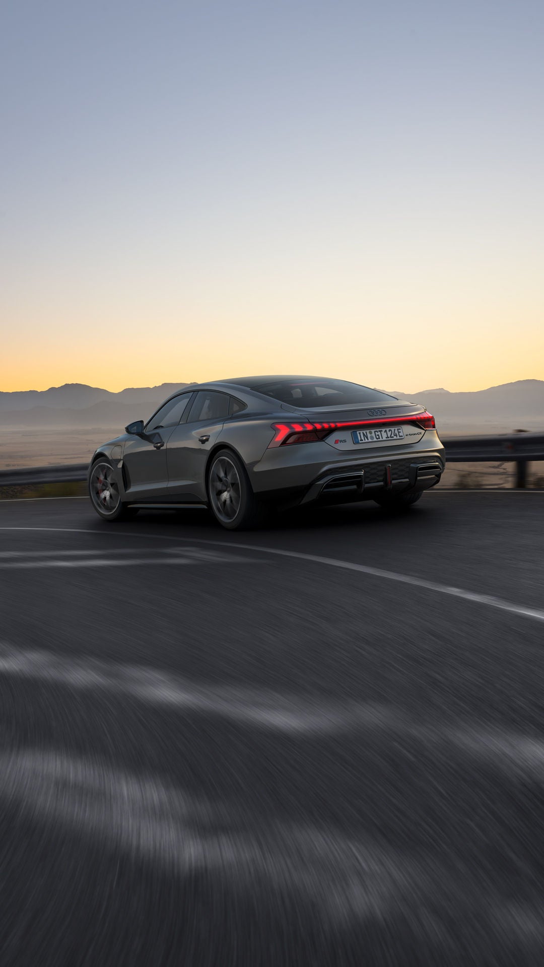 Ein grauer Audi RS e-tron GT fährt auf einer kurvigen Bergstraße bei Sonnenuntergang, mit leuchtenden Rücklichtern.