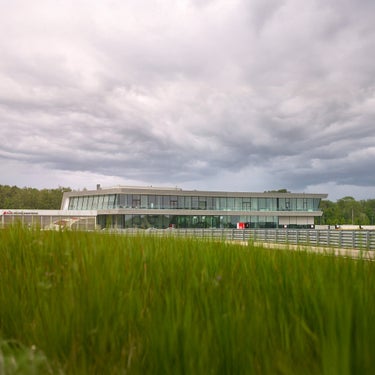 Blick über eine grüne Wiese auf die Rennstrecke Neuburg. Im Hintergrund ist das Gebäude des Audi driving centers zu sehen.