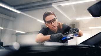 Mitarbeiterin mit Schutzbrille und Handschuhen repariert die Windschutzscheibe von einem Fahrzeug in einer Werkstatt.