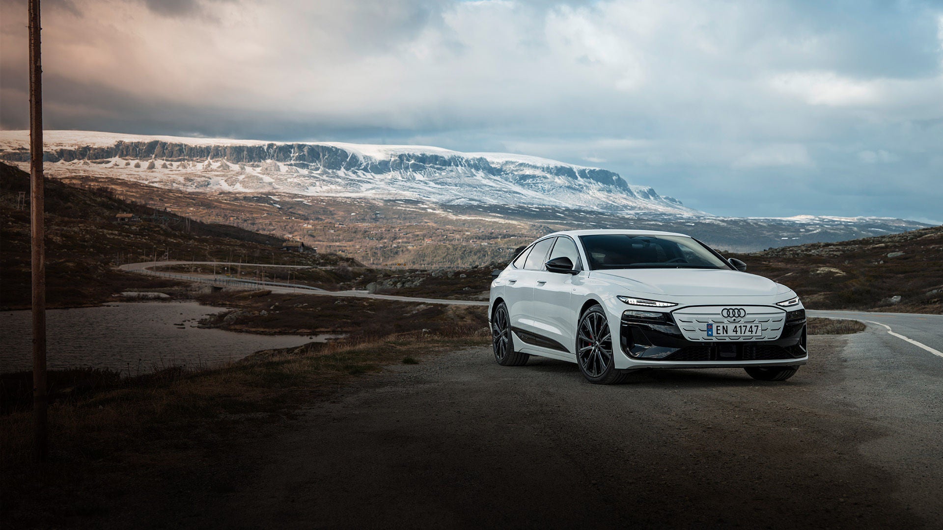 Weißer Audi A6 Sportback e-tron vor einer skandinavischen Naturkulisse mit Nadelwäldern, schneebedeckten Bergen und einem Flusslauf.