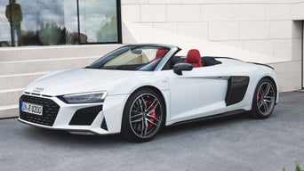 Weißer Audi R8 Spyder mit offenem Verdeck und rotem Interieur vor hellem Gebäude im Stand.