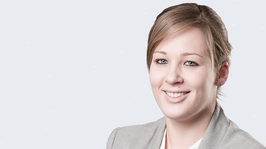 Marlen Muhr: Porträt einer lächelnden Frau mit blondem Haar zum Zopf im grauen Blazer mit weißem Oberteil vor hellgrauem Hintergrund.