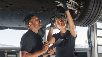 Zwei Audi-Mitarbeitende inspizieren gemeinsam die Fahrzeugunterseite in einer hellen Werkstattumgebung.