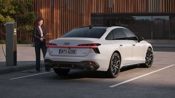Die A6 Limousine e-hybrid in weiß steht auf einem Parkplatz vor einer Gebäudefassade. Eine junge Frau schließt die A6 Limousine e-hybrid an eine Ladestation an.