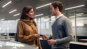 Zwei Personen übergeben sich einen Autoschlüssel in einem hellen Innenraum mit Glaswänden und Deckenleuchten.