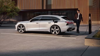 Ein weißer Audi A6 Avant e-hybrid lädt an Ladesäule auf einem Parkplatz, junger Mann steht lächelnd daneben, modernes Gebäude im Hintergrund.