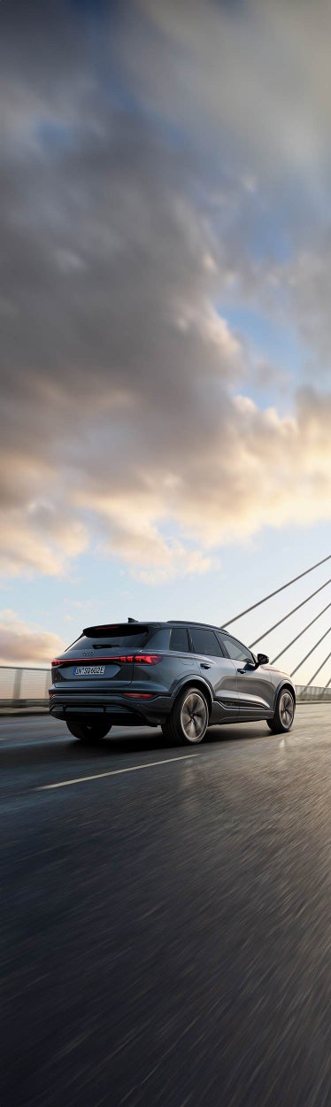 Grauer Audi SQ6 SUV e-tron fährt über eine Brücke bei Sonnenaufgang, mit Seilverspannung im Hintergrund.