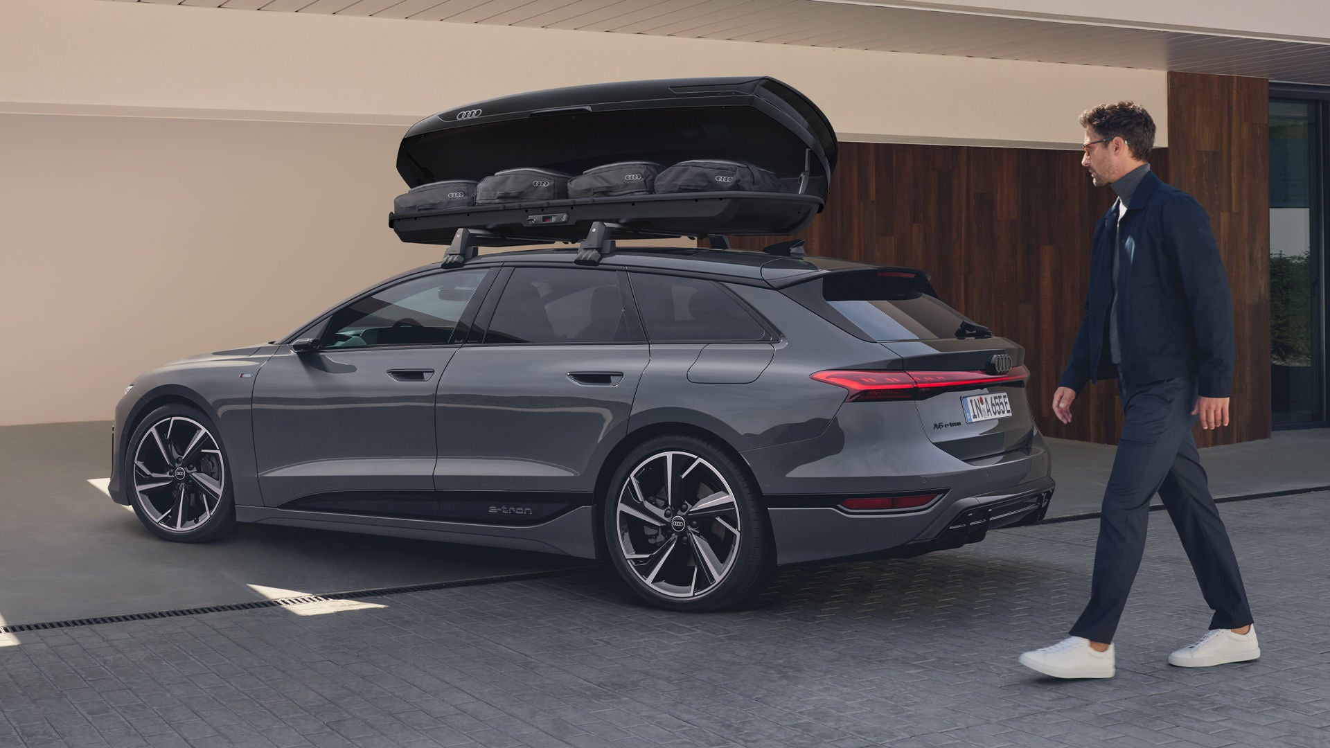 Der A6 Avant e-tron steht schräg vor einem modernen Haus, ein Mann bewegt sich auf das Fahrzeug zu. Auf dem Dach ist eine Dachbox montiert, deren Klappe geöffnet ist. Taschen sind in der Dachbox zu sehen.