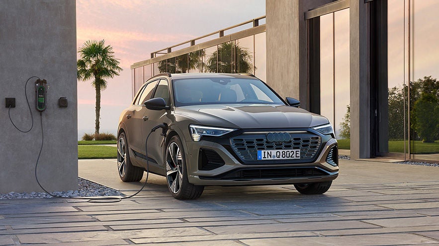 Grauer Audi Q8 e-tron wird vor einem modernen Gebäude mit Palmen im Hintergrund an einer Wallbox geladen.