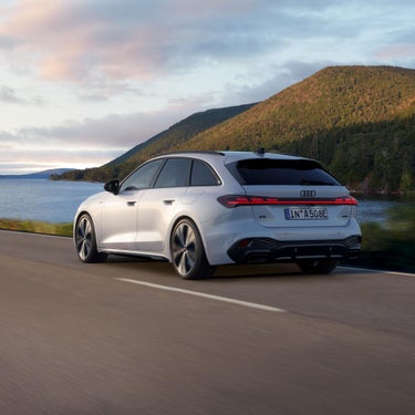 Ein weißer Audi A5 Avant e-hybrid, von schräg hinten zu sehen, fährt auf einer Straße an einem See entlang. Im Hintergrund sind Hügel zu erkennen.