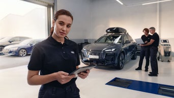 Eine Frau mit einem Tablet in der Hand in einem Autohaus im Hintergrund steht ein Audi und zwei Leute mit einem Laptop.