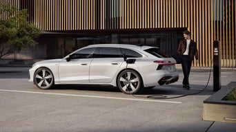 Der A6 Avant e-hybrid steht seitlich vor dem Eingangsbereich eines großen Gebäudes und wird über ein Stromkabel geladen. Am Heck des Fahrzeugs steht ein junger Mann.