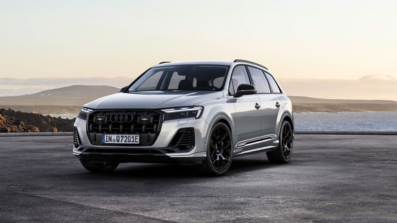 Ein weißer Audi Q7 TFSI e mit schwarzem Kühlergrill und schwarzen Felgen steht vor einer malerischen Küstenlandschaft mit sanften Hügeln im Hintergrund, während die Abendsonne den Himmel in warme Farben taucht.