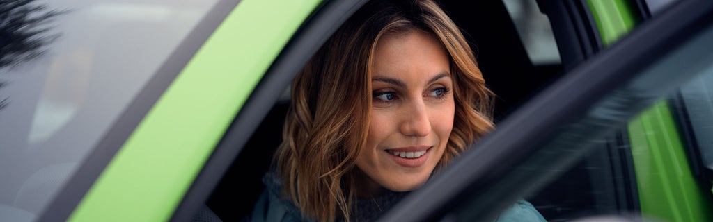 Woman with brunette hair looks out of the open driver's door of a light green car with a smile.