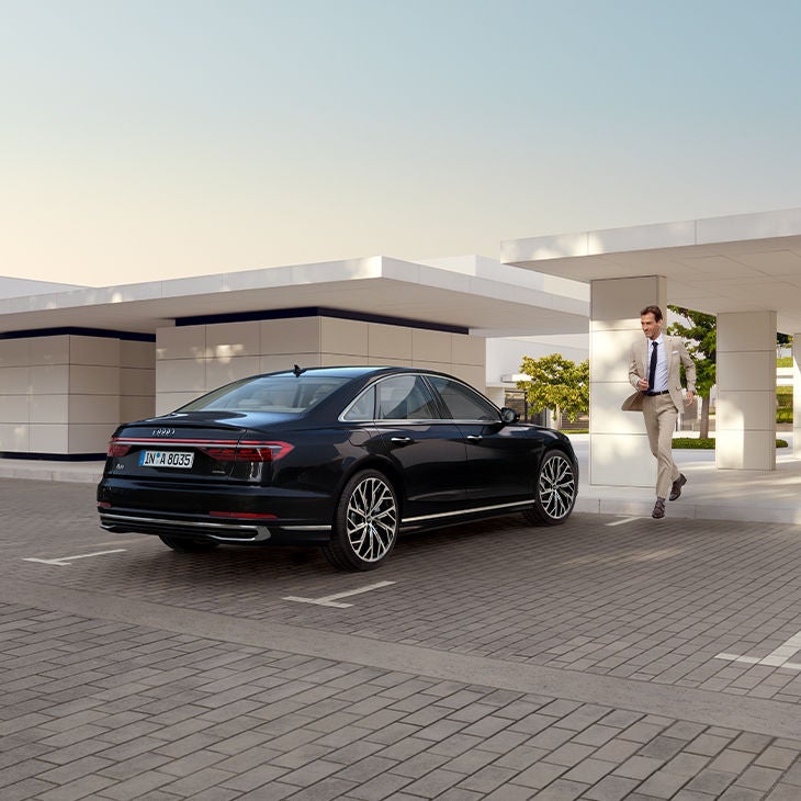 Ein dunkler Audi A8 parkt vor einem weißen modernen Haus. Ein Mann im Anzug rennt auf das Auto zu.