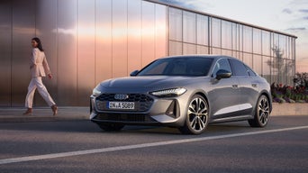 Eine Frau geht an einer silbernen Audi A5 Limousine vorbei, der vor einem modernen Glasgebäude parkt.