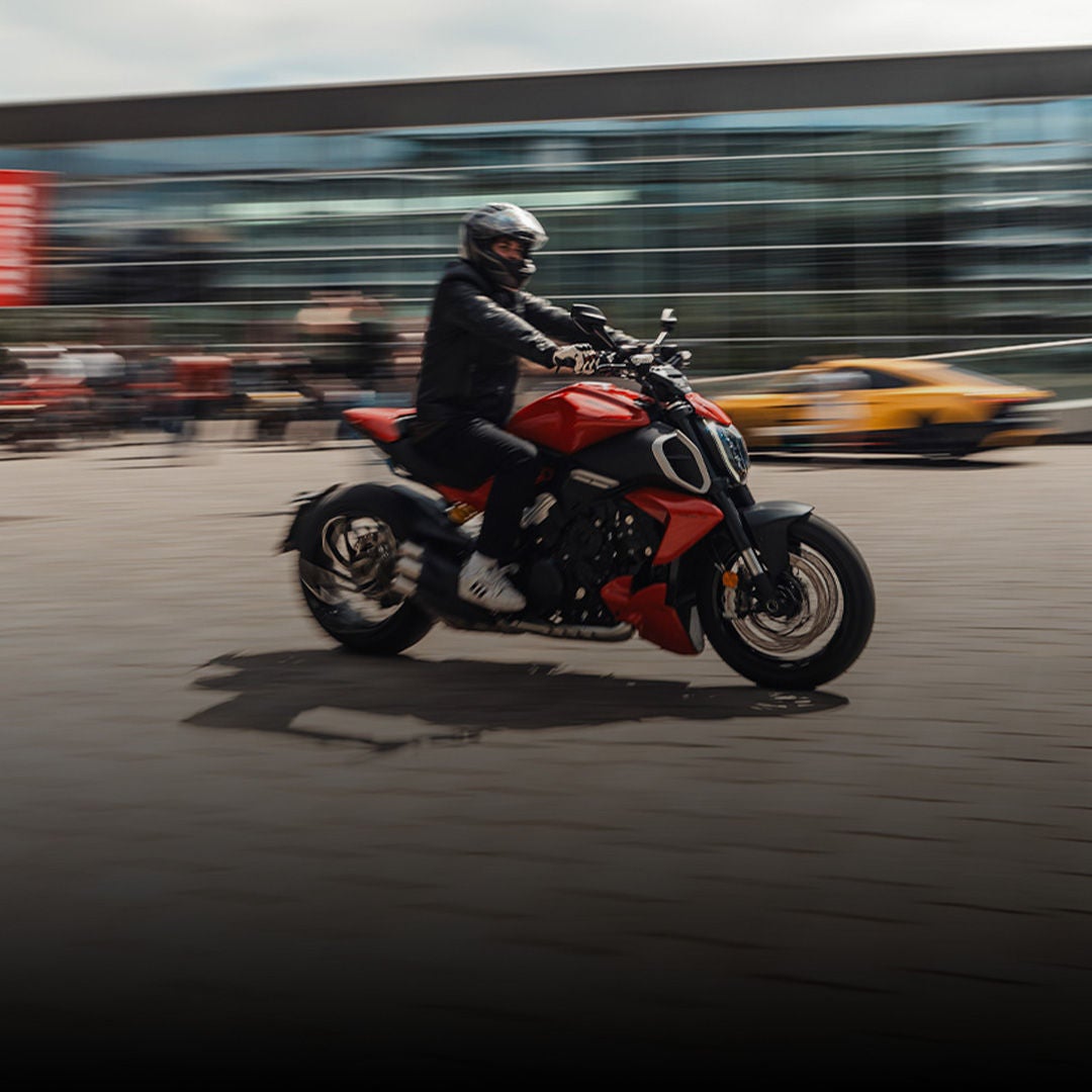 Motorradfahrer fährt dynamisch über Platz vor modernem Gebäude, Hintergrund mit Bewegungsunschärfe.