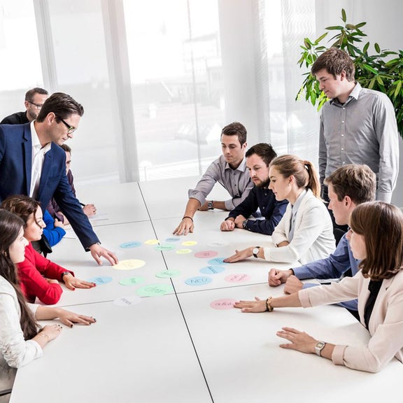 Gruppe sitzt an weißen Tischen in einem modernen Konferenzraum und arbeitet gemeinsam mit bunten Moderationskarten.