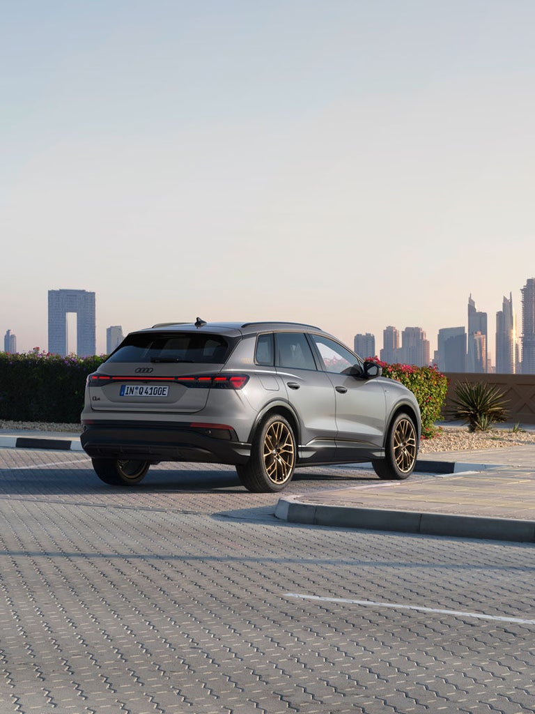 Heckansicht eines grauen Audi Q4 e-tron mit goldenen Felgen, Skyline und blühender Hecke bei Sonnenaufgang, geparkt auf Pflasterfläche.