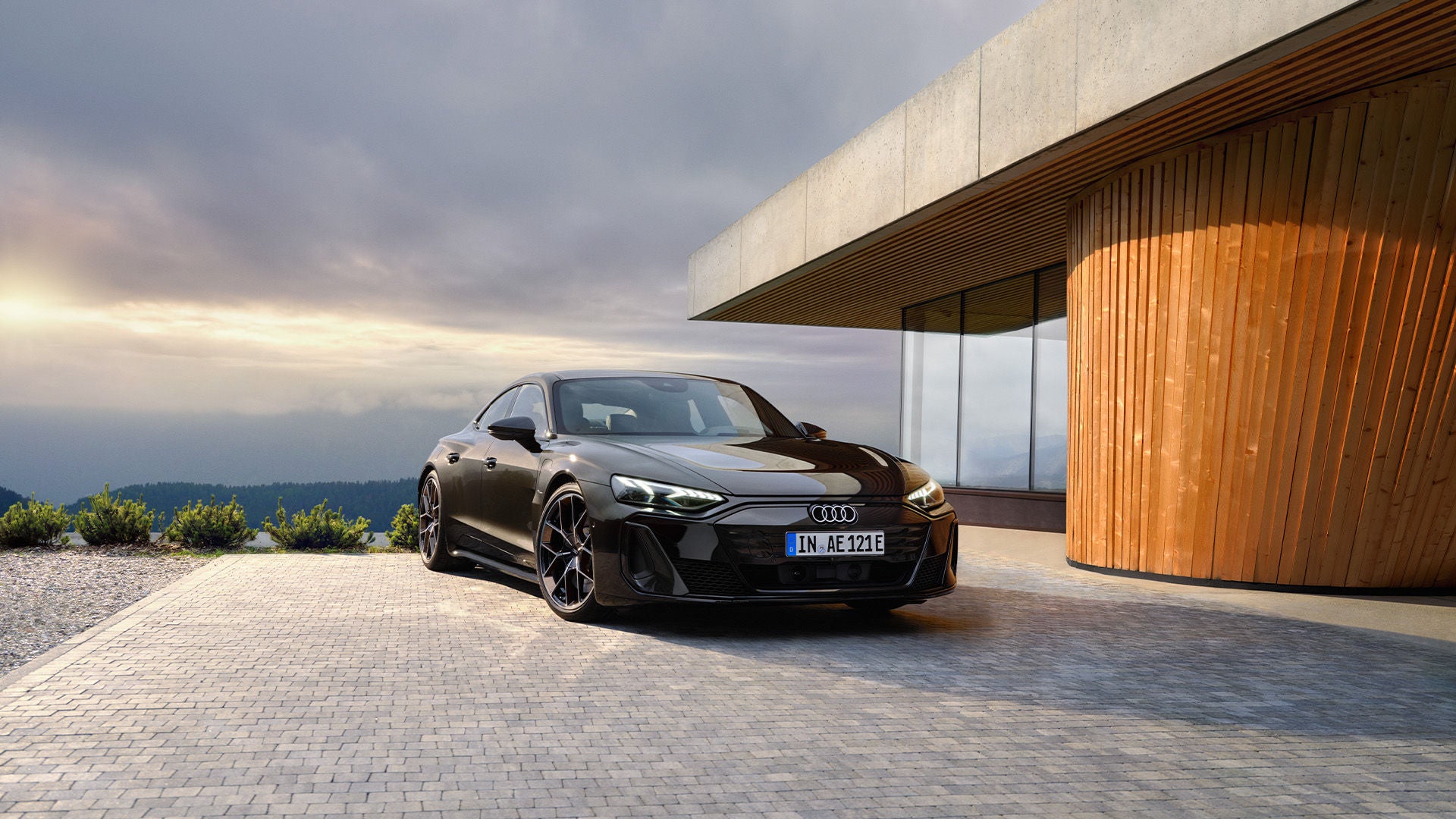 Schwarzer Audi e-tron GT parkt vor einem modernen Haus mit Holzfassade bei bewölktem Himmel.