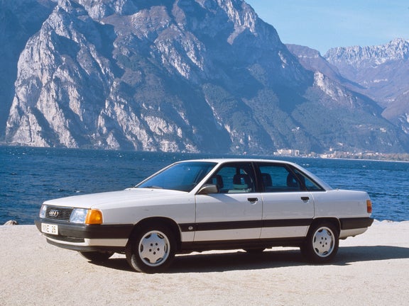 A white Audi 100 TDI in the front side view from the left in front of a mountain backdrop with a mountain lake