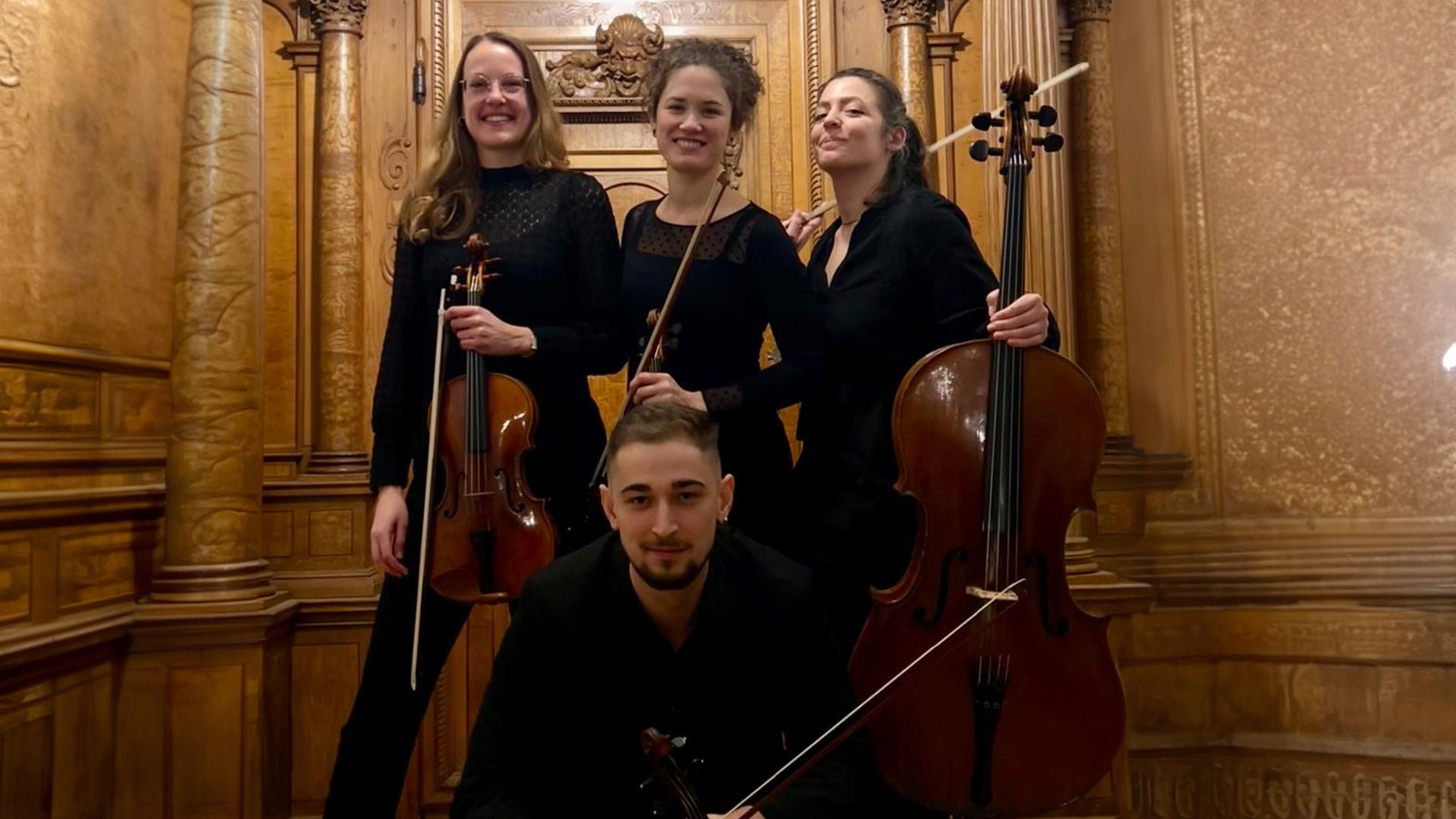 Aeterna Quartett posiert vor einer hölzernen Wand mit Instrumenten in den Häden (3 Frauen hinten stehend und ein Mann, vorne sitzen, in schwarzen Outfits). 