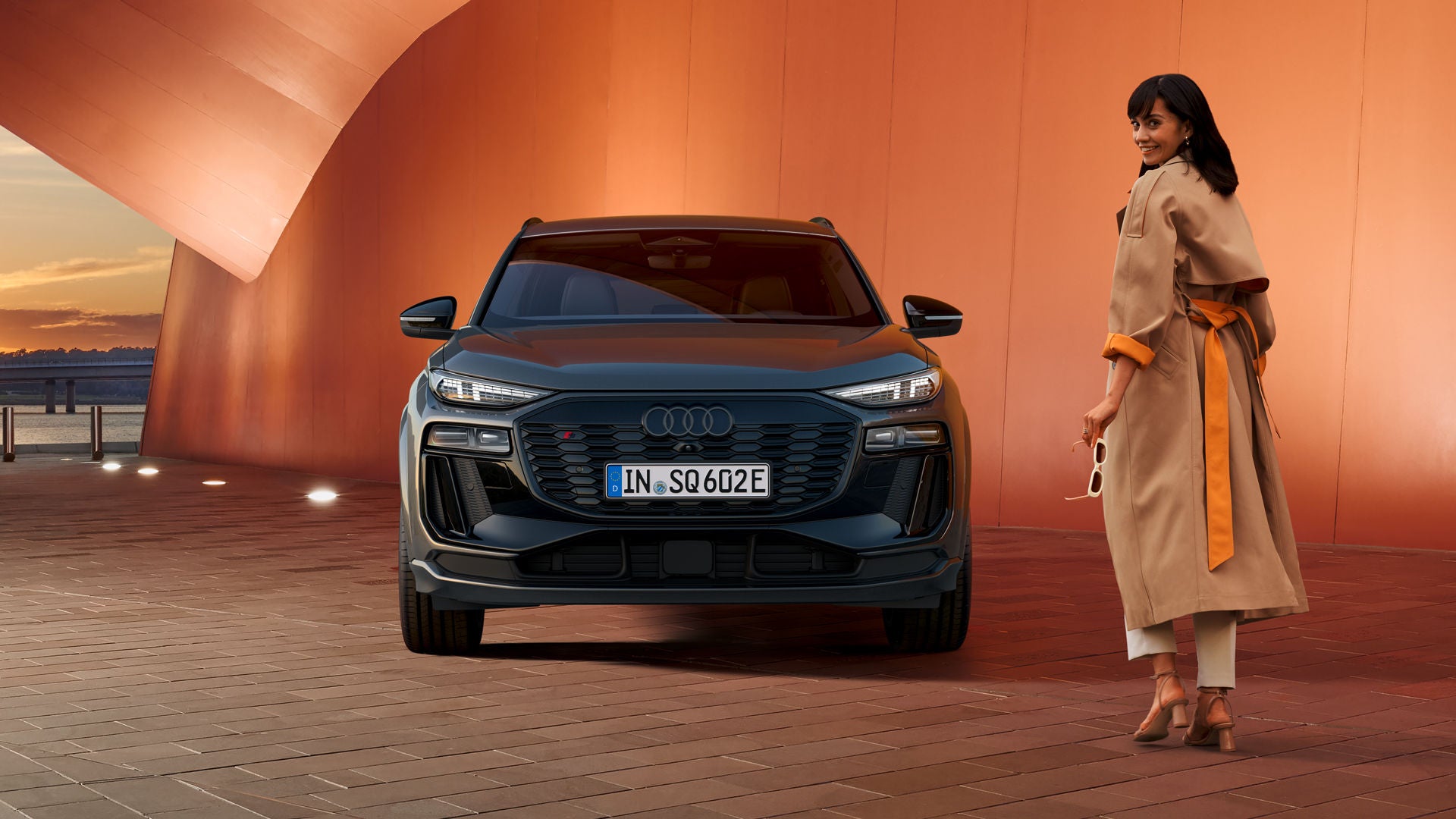 Seitliche Front des SQ6 SUV e-tron, welcher neben einer orangenen Gebäudefassade an eine Ladestation angeschlossen ist. Im Hintergrund ist das Meer, sowie die Skyline einer Stadt zu erkennen.