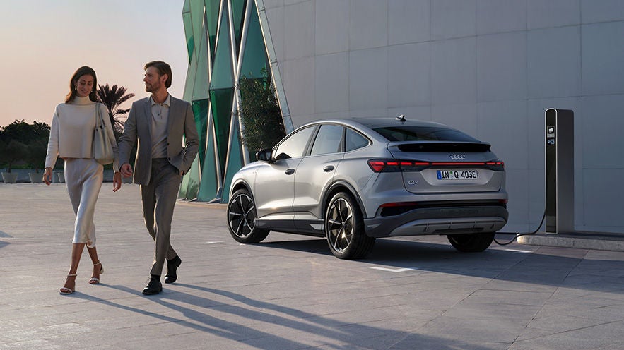 Ein silberner Audi Q4 e-tron lädt an einer Ladesäule, während ein Paar elegant gekleidet daran vorbeigeht.