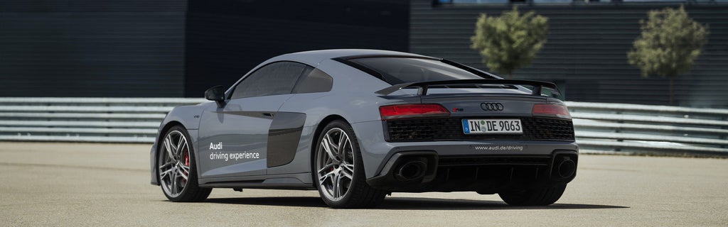 Mattgrauer Audi R8 Coupé V10 performance quattro mit weißer 'Audi driving experience' Aufschrift auf Teststrecke, Heckansicht mit Spoiler, Leitplanken im Hintergrund.