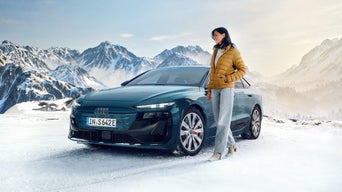 Frau in gelber Jacke steht neben blauem Audi auf verschneiter Fläche vor Bergkulisse.