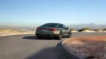 A green Audi car maneuvers around a coastal bend, showcasing its performance against a backdrop of mountains and blue skies.