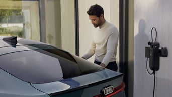 A young man stands at the rear of the S6 Sportback e-tron and connects the vehicle to a charging station mounted on the wall.