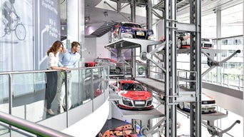 Eine Frau und ein Mann stehen am Geländer des Audi museum mobile in der zweiten Etage und blicken auf den Paternostra mit verschiedenen Audi Modellen.