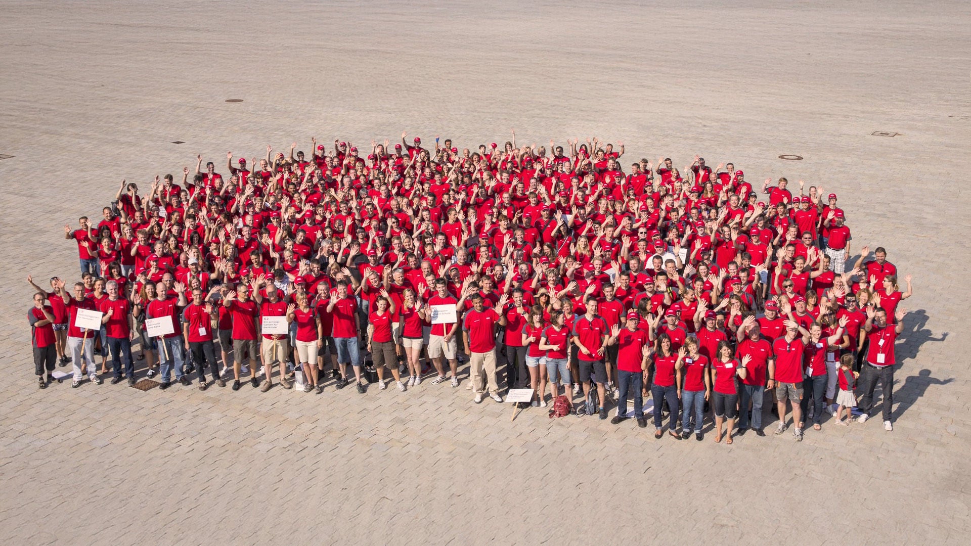 Aerial view of a large group of Audi employees wearing red shirts. They are waving at the camera.
