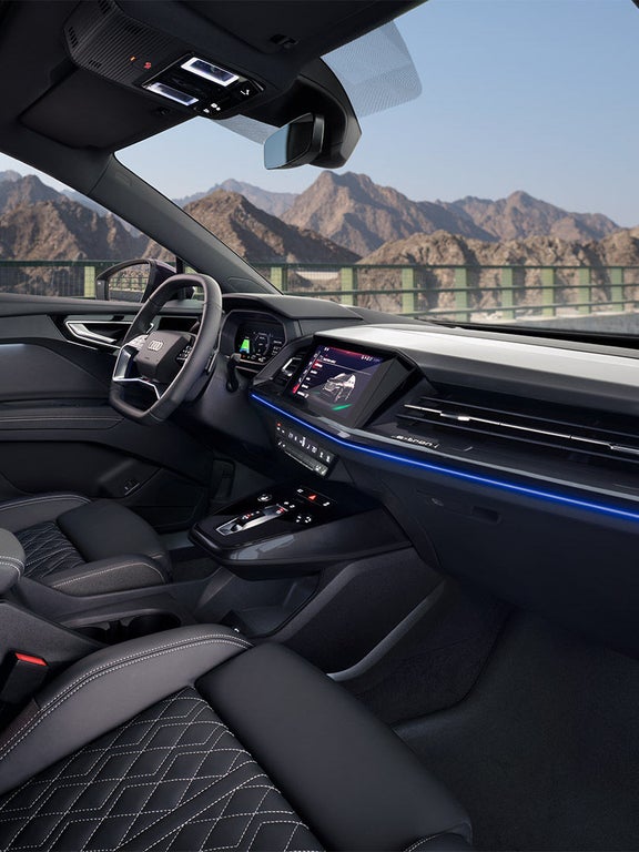 Blick in den Innenraum eines Audi Q4 e-tron mit Digitaldisplay, blauer Ambientebeleuchtung und Berglandschaft sowie einem Paar draußen.
