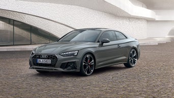 Grauer Audi A5 Coupé auf Kopfsteinpflaster vor moderner Architektur mit geschwungenen weißen Fassaden.