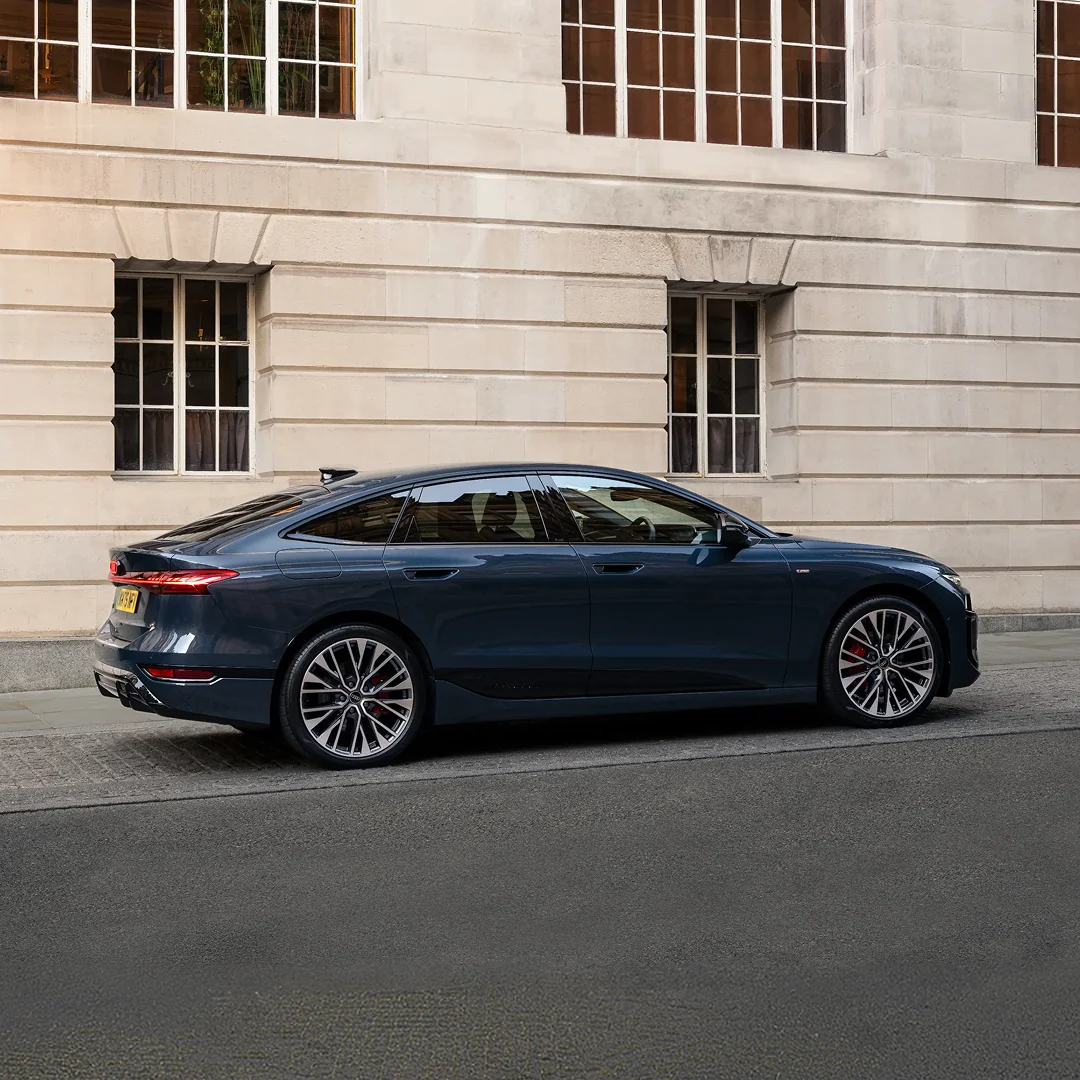 Dark blue Audi e-tron Sportback parked on a city street, shown in full side profile against a stone building facade. The vehicle features a sleek fastback roofline, flush door handles, large multi-spoke alloy wheels and slim rear LED lights. Daylight reflects across the sculpted bodywork, highlighting the aerodynamic design and premium exterior styling.